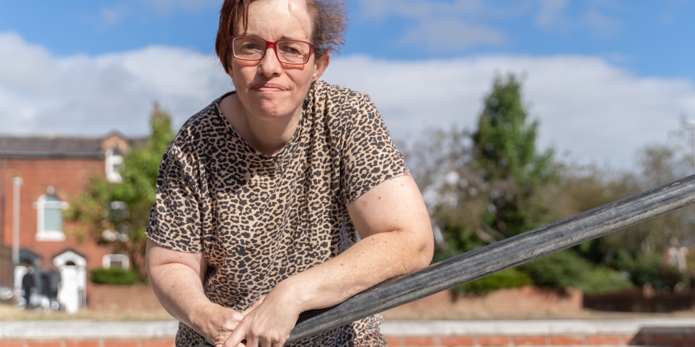 Louise is learning against a rail looking at the camera, she's wearing a leopard print top. Behind her are red brick buildings, some stairs and blue sky.