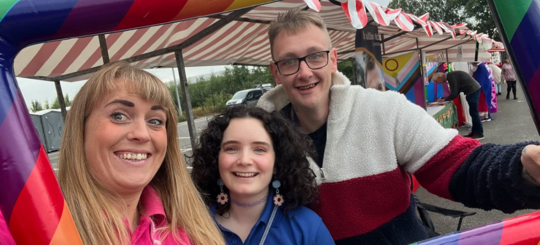 Image shows three members of the Healthwatch Cumberland team posing for a photo with an inflatable rainbow photo frame held around their faces.