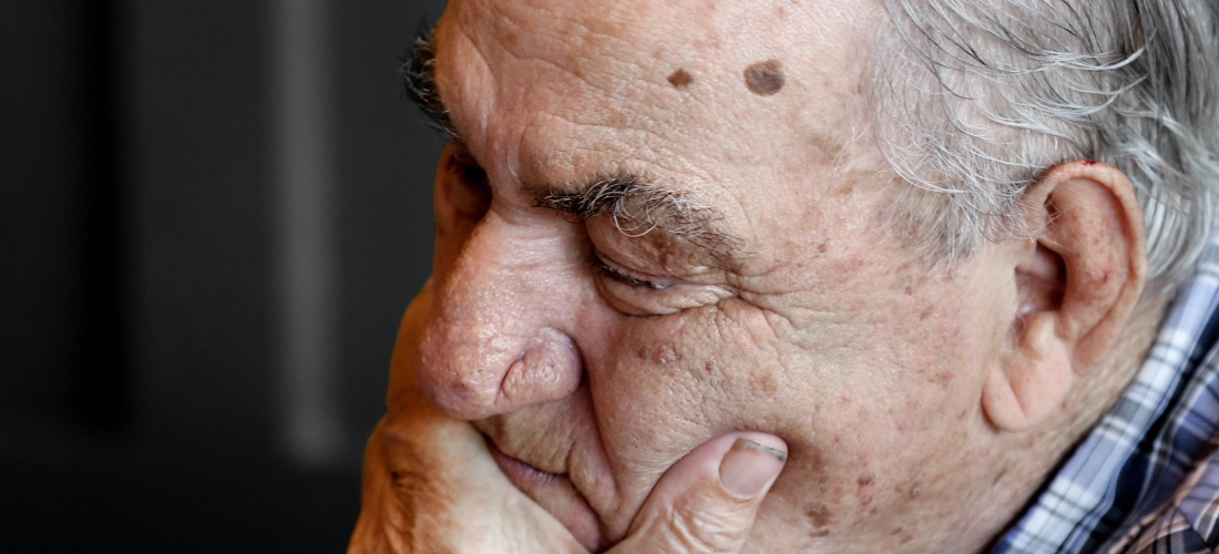 An old gentleman sat with his hand covering the bottom part of his face, he looks worried.